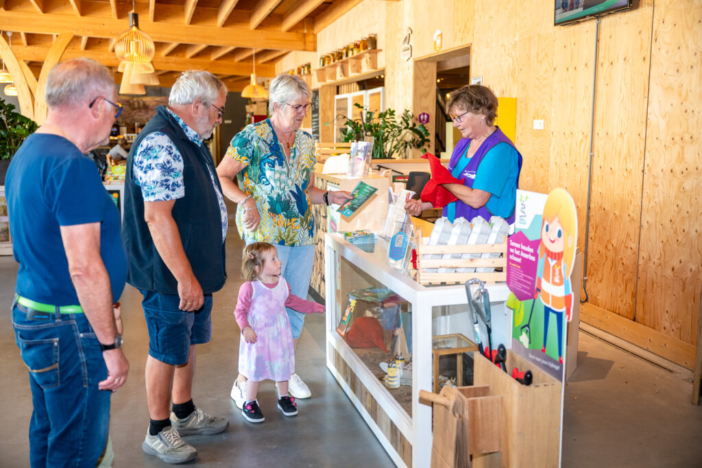Balievrijwilliger staat achter de balie en geeft iets aan bezoekers voor een activiteit.