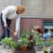 Een man met een schept aarde in een geveltuin. Vrouw plant een plant in de aarde. Er staan verschillende planten in potjes die nog gepland moeten worden.