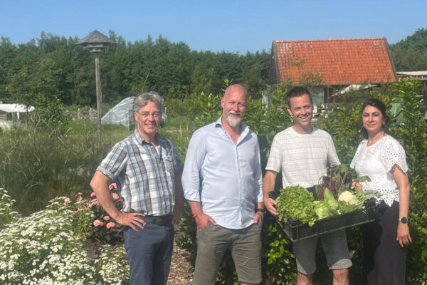 Vier mensen naast elkaar in een veld. De lucht is blauw. De derde persoon heeft een krat met groenten vast.