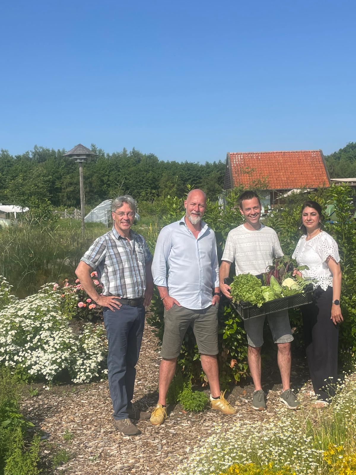 Vier mensen naast elkaar in een veld. De lucht is blauw. De derde persoon heeft een krat met groenten vast.