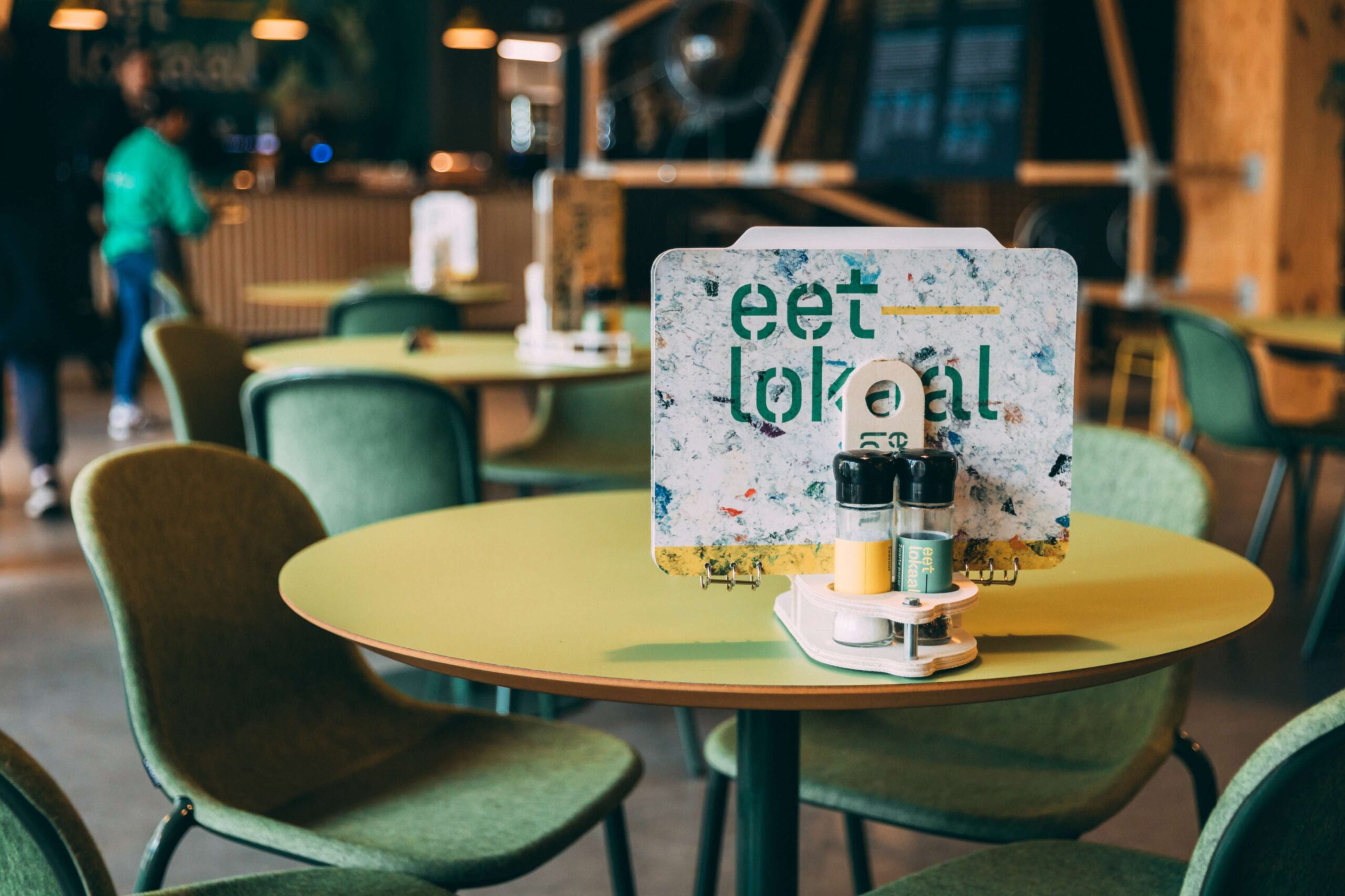Groene ronde tafel en stoelen met daarop een menukaart en peper- en zoutstel in Het Eetlokaal, het restaurant van het Duurzaamheidscentrum Assen